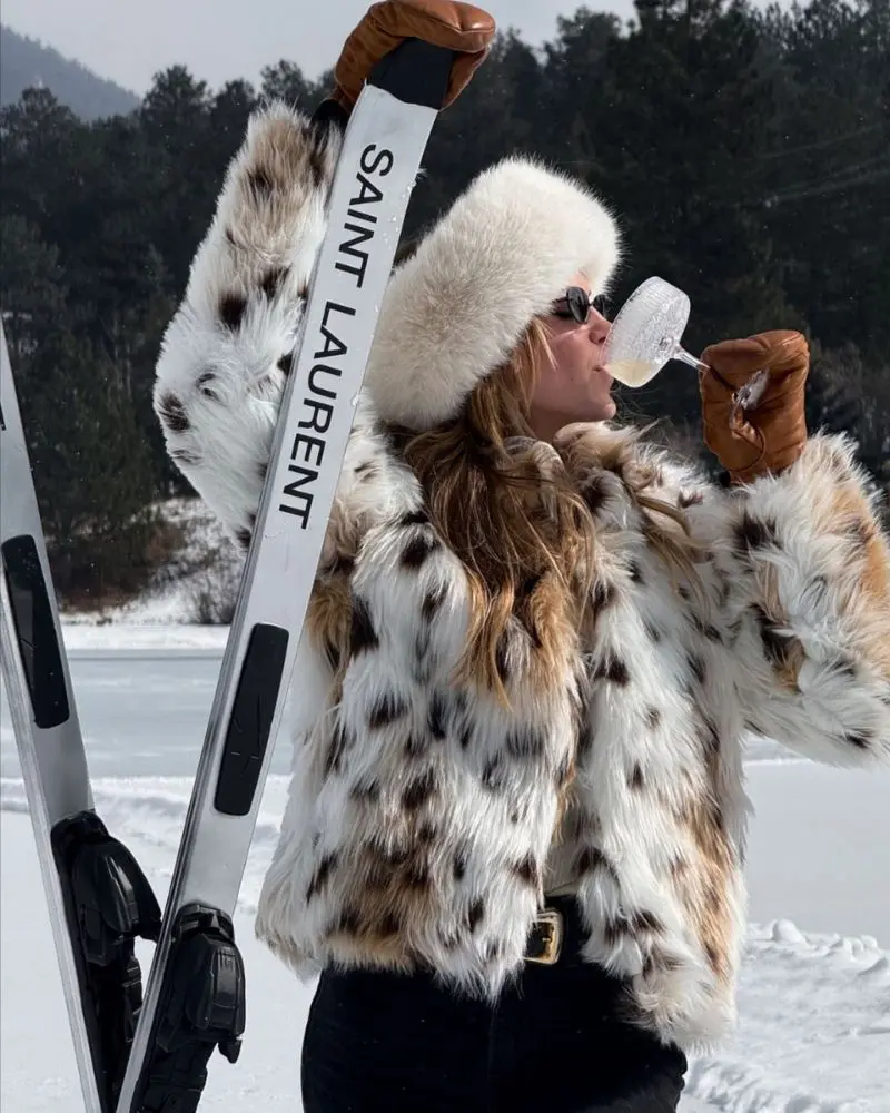 Mujer con abrigo y gorro invernal brindando con copa junto a unos esquís sobre la nieve (après-ski)